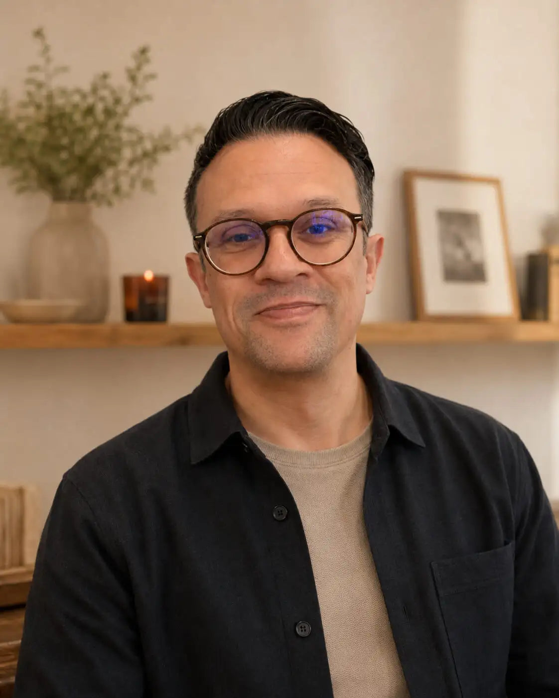 Man wearing dark brown framed glasses and a black button-up shirt over a beige undershirt